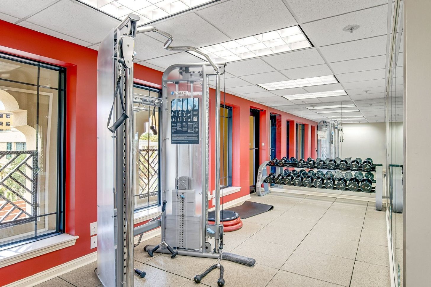 Un gimnasio con equipo de ejercicio, un soporte de mancuernas en la pared y una ventana con vistas a edificios.
