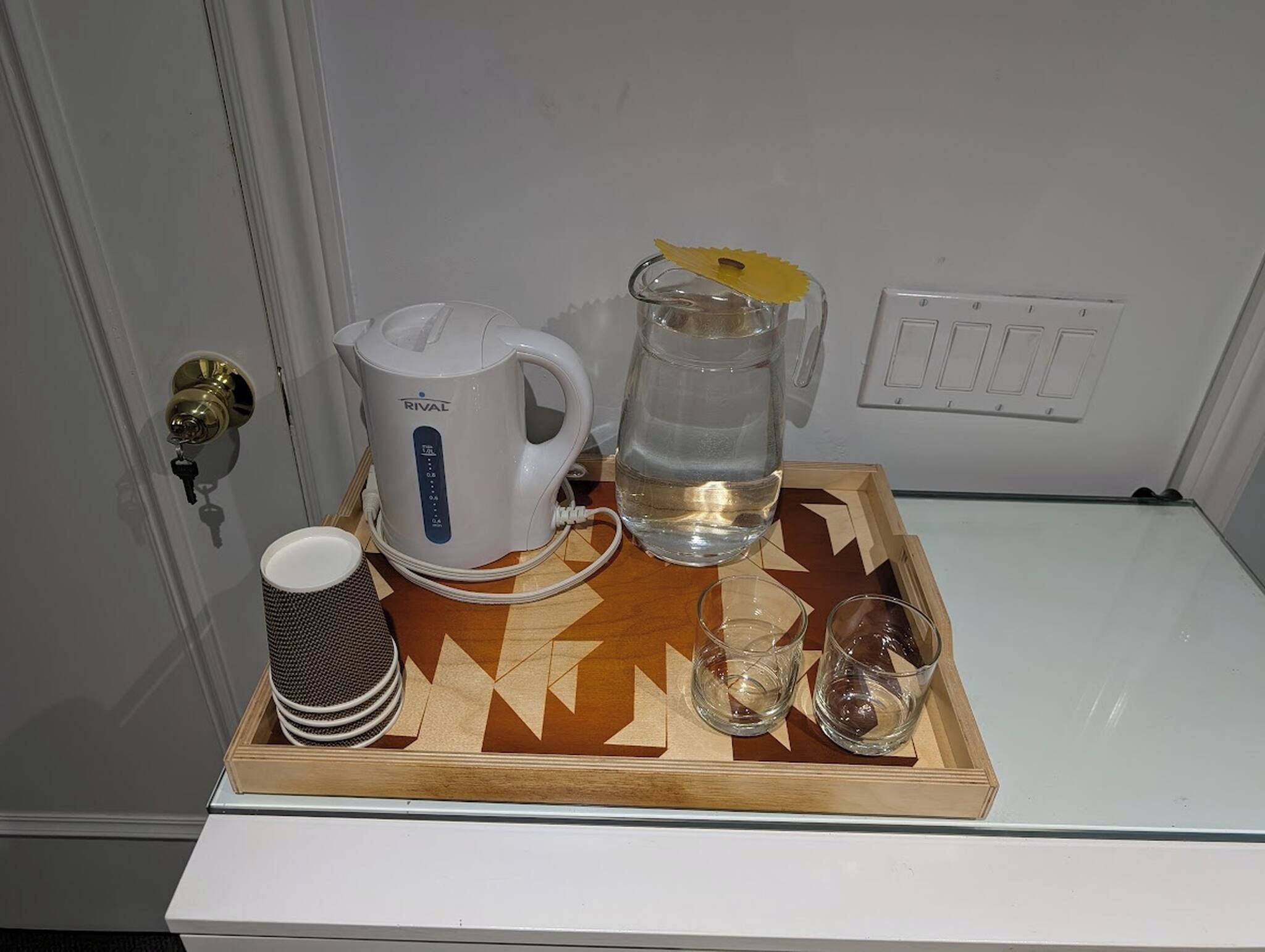 A tray with a white electric kettle, a glass pitcher, a cup, and glasses on a wooden tray.
