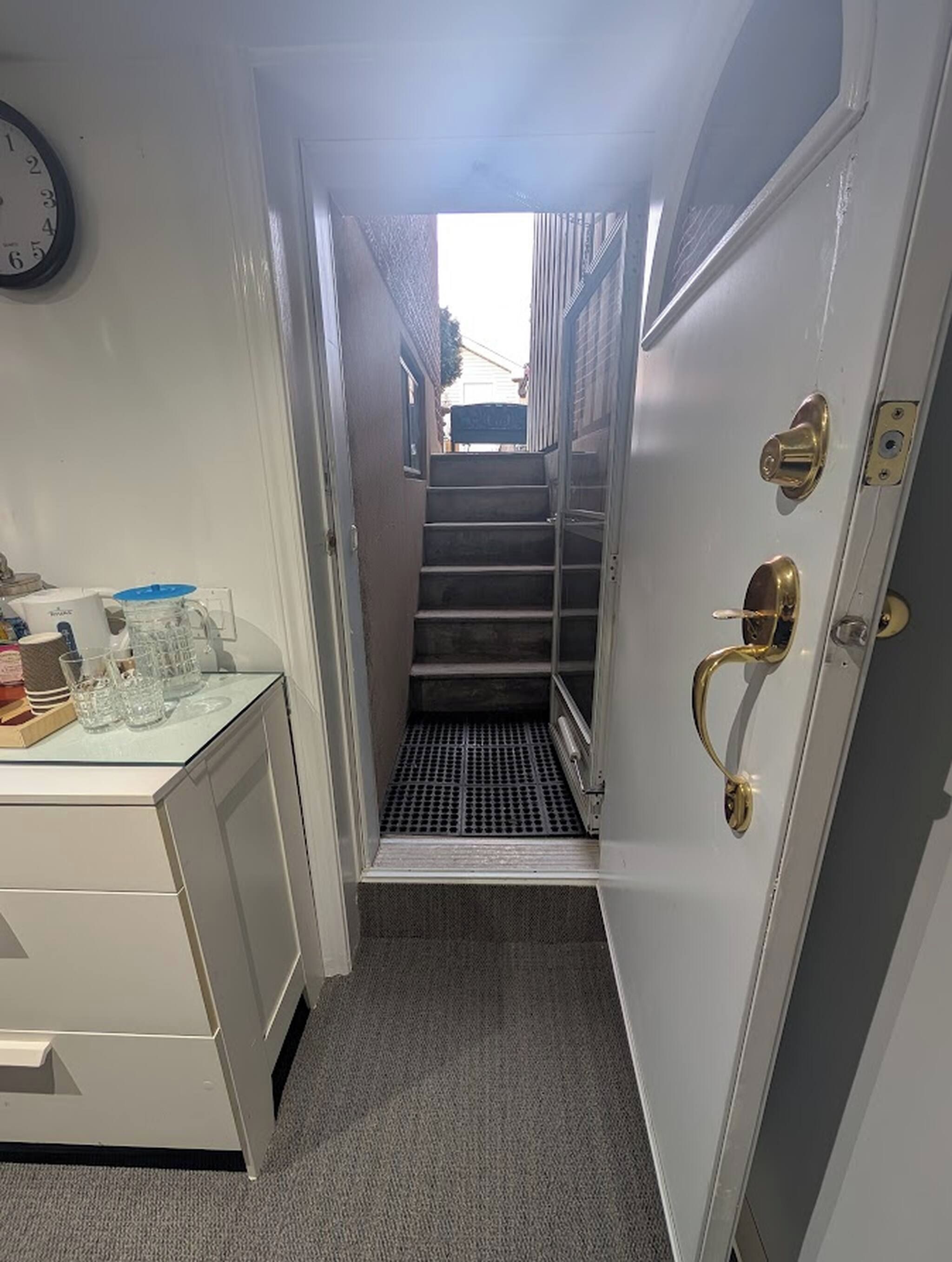 A narrow hallway with a staircase leading to an upper level, a door with a brass handle, and a clock on the wall.