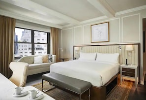 Quarto de hotel com uma cama grande, um sofá, uma escrivaninha com cadeira, uma mesa de centro e uma janela com cortinas.