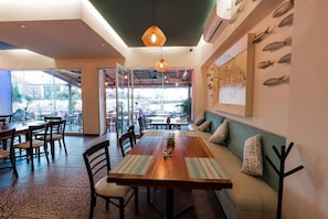 Un comedor interior con mesas, sillas y banco de madera. Hay luces colgantes, un mural con motivos marinos en la pared y vistas a través de puertas de vidrio.