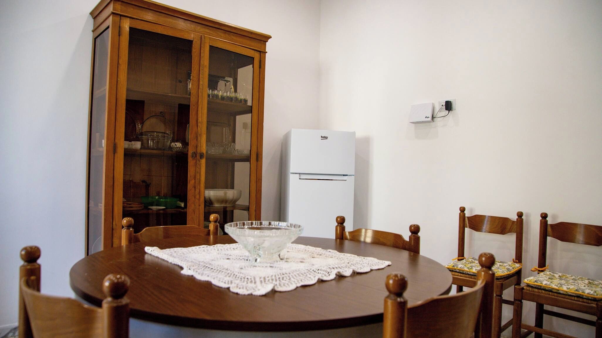 A dining area with a round wooden table, six wooden chairs with cushioned seats, a white lace tablecloth, a glass bowl, and a white refrigerator.
