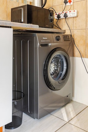 Een wasmachine met een voorlaadopening, een bedieningspaneel, een stopcontact en een prullenbak in een keuken.