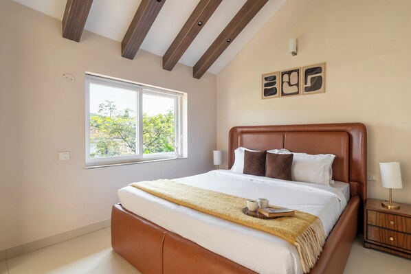 A hotel room with a large bed, a window with a view of trees, a bedside table with a lamp, and a wall-mounted artwork.