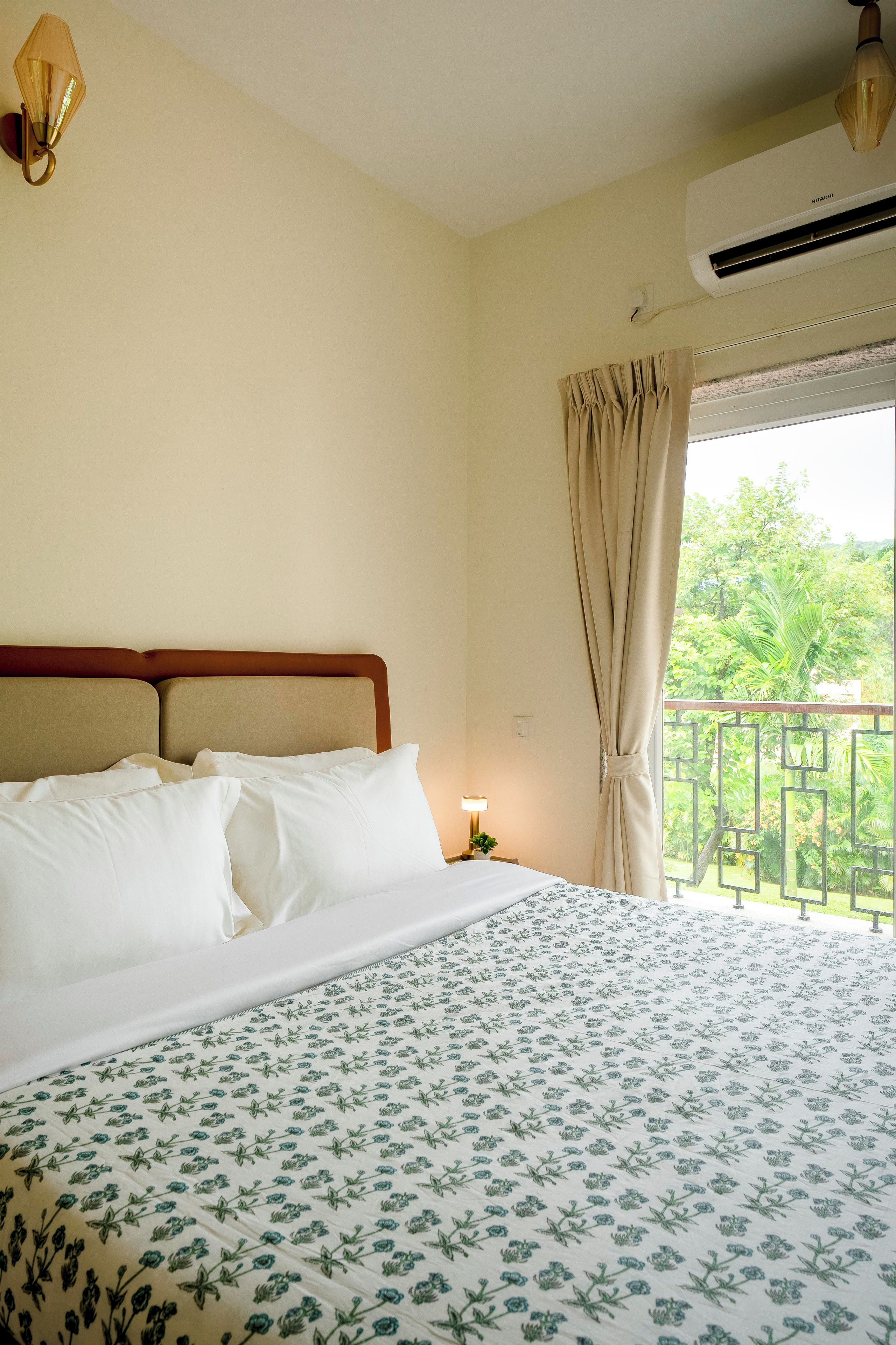 A hotel room with a bed, a nightstand, a lamp, a window with curtains, and an air conditioning unit.