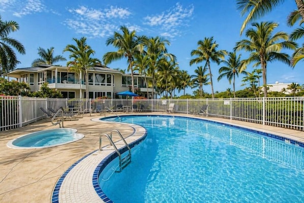 Ein Swimmingpool mit einer gefliesten Kante, ein Liegebereich mit Stühlen und ein Gebäude mit einem Balkon.