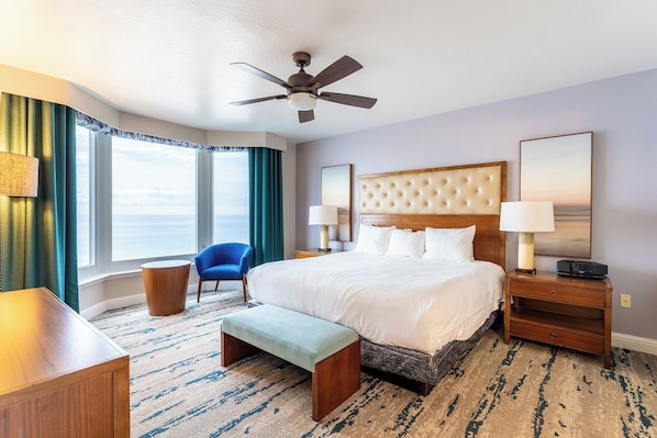 A hotel room with a large bed, a ceiling fan, a view of the ocean, a blue chair, and a wooden nightstand with a lamp. - Panama City Beach | Family Coast Base | Pool + Hot Tub | 2BR Condo (Panama City)