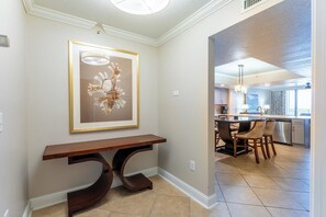 A hallway leading to a dining area with a wooden bench, a framed artwork, and a view into a kitchen with modern appliances and a dining table. - Panama City Beach | Family Coast Base | Pool + Hot Tub | 2BR Condo (Panama City)