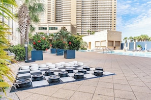 A pool area with a checkered pattern of tires and potted plants, a poolside lounge, and a tall building in the background. - Panama City Beach | Family Coast Base | Pool + Hot Tub | 2BR Condo (Panama City)