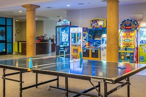 Uma mesa de tênis de mesa com o logotipo Brunswick, rodeada por jogos de fliperama incluindo 'Safecracker' e 'Piratas'.