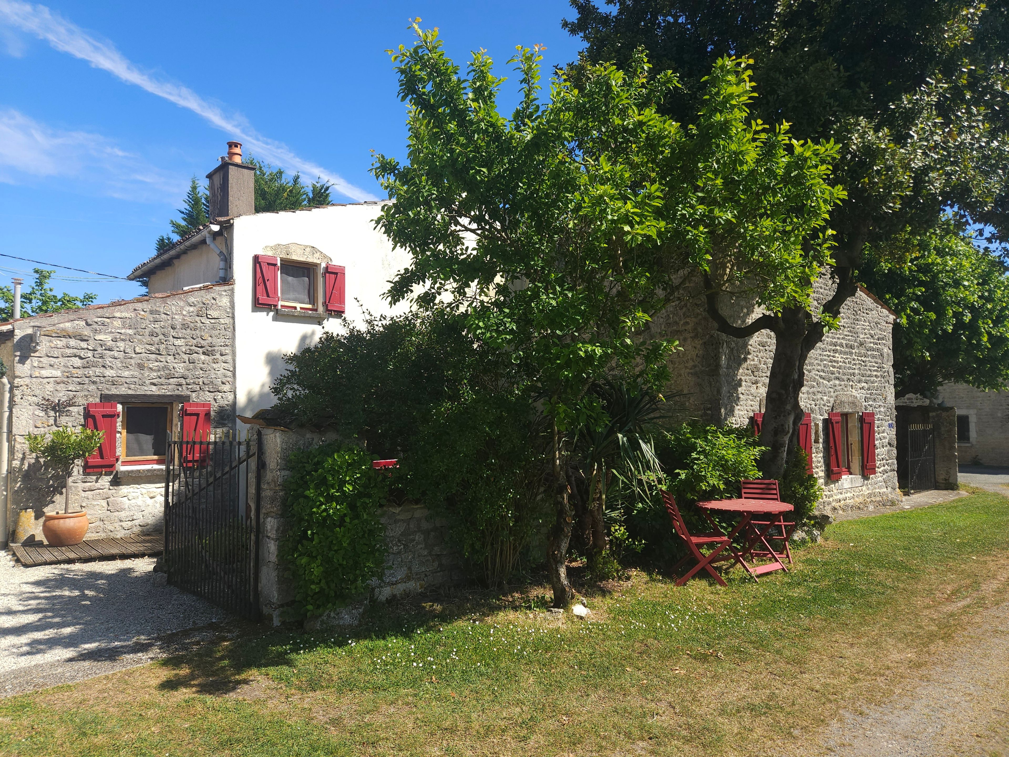 Une maison en pierre avec des volets rouges, un petit jardin, ainsi qu’une table et des chaises rouges.