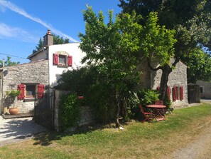 Une maison en pierre avec des volets rouges, un petit jardin, ainsi qu’une table et des chaises rouges.