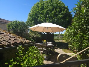 Un espace extérieur aménagé avec une table et des chaises, protégé par un grand parasol, et entouré de verdure et d’un arbre.