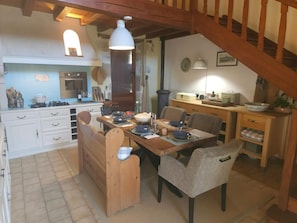 Een gezellig eetgedeelte met een houten tafel voor vier personen, een keuken met fornuis en kastjes, en een trap naar een hoger niveau.