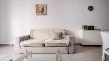 A minimalist living room with a sofa, a glass coffee table, a wall-mounted clock, and a bookshelf.
