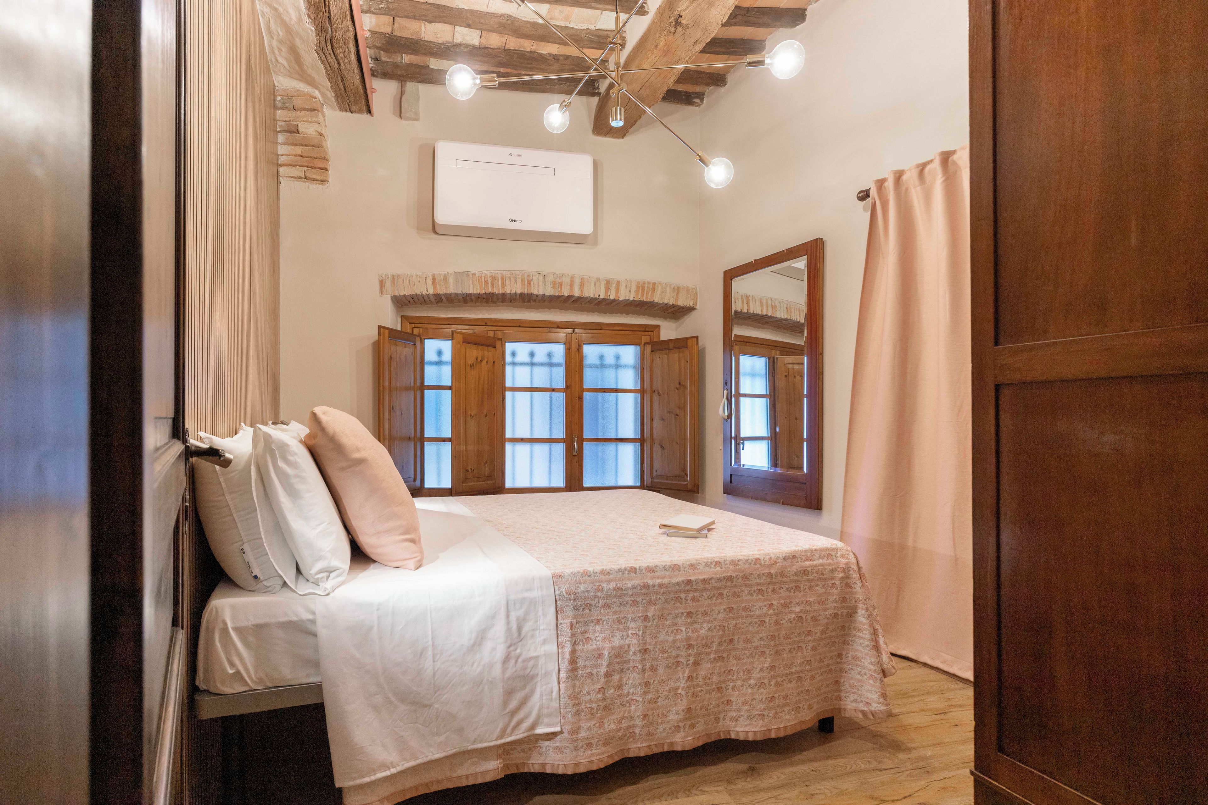 Een slaapkamer met een bed, een raam met houten luiken, een plafondlamp en een spiegel aan de muur.
