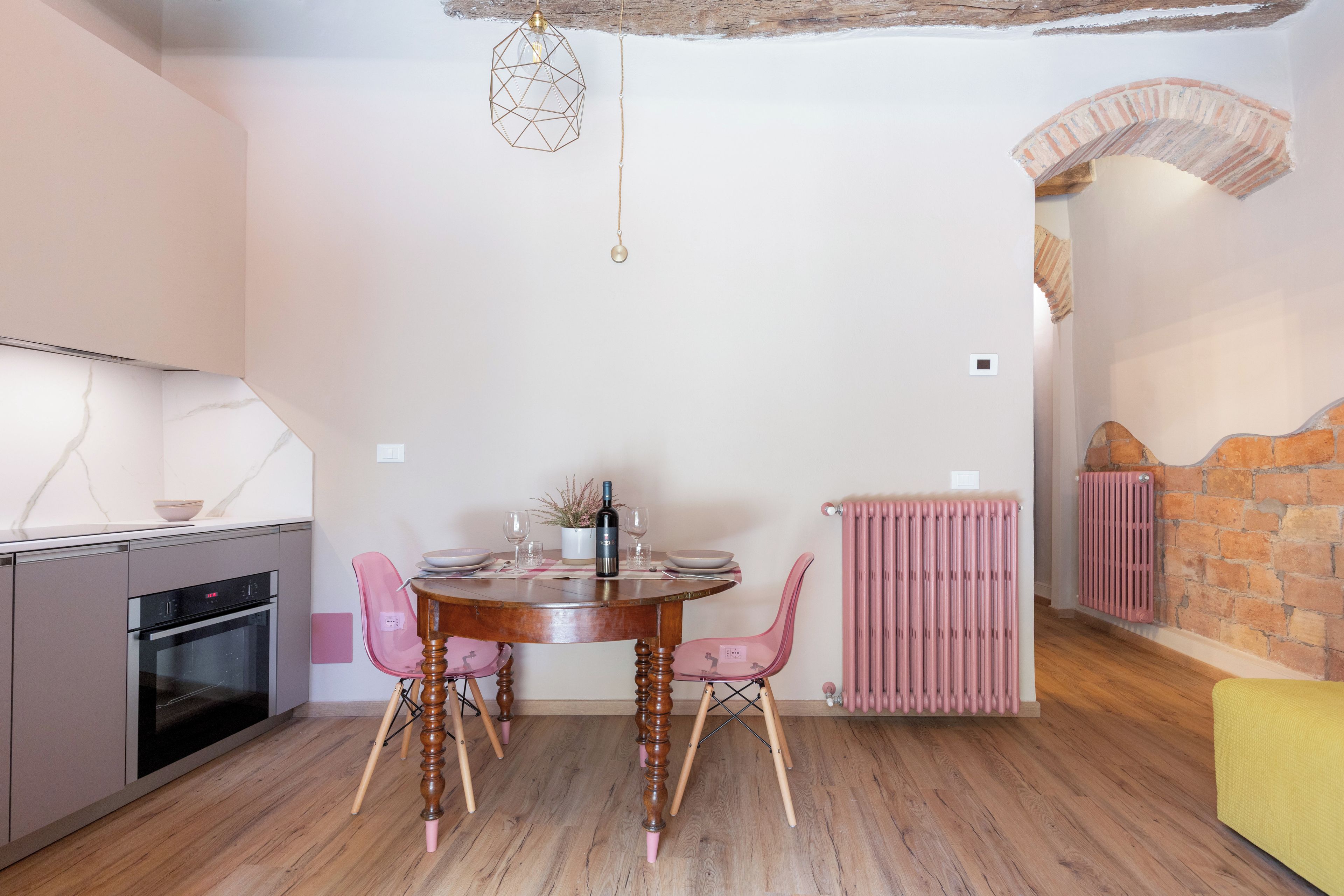 Een eetgedeelte met een ronde tafel, roze stoelen, een wijnfles en een hanglamp.
