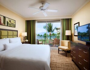 Habitación de hotel con cama, escritorio, silla, televisión y balcón con vistas a palmeras y edificios.