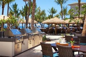 An outdoor dining area with grill stations, wicker furniture, and palm trees. - Maui | Couples Island Weekend | Pool + Beach (Lahaina)