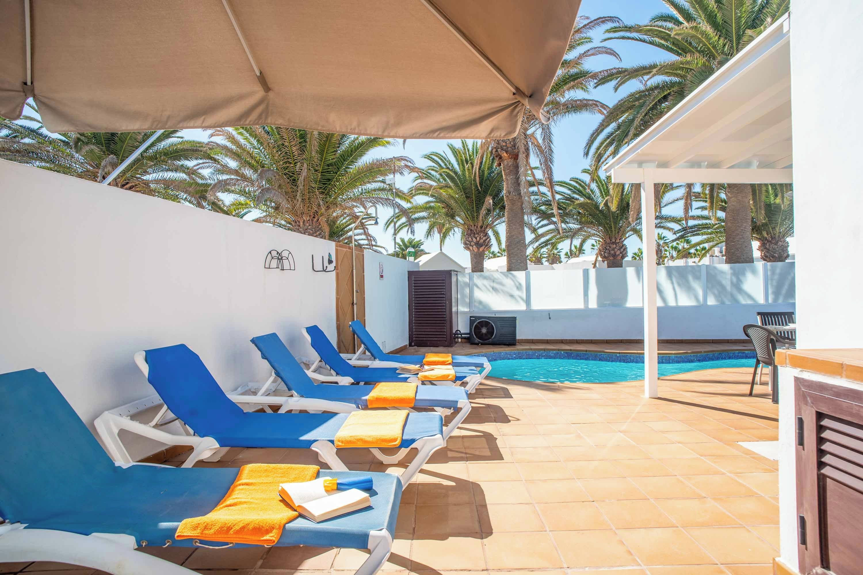 Un espace aménagé au bord de la piscine, avec des chaises longues bleues et des serviettes orange, une piscine et des palmiers en arrière-plan.