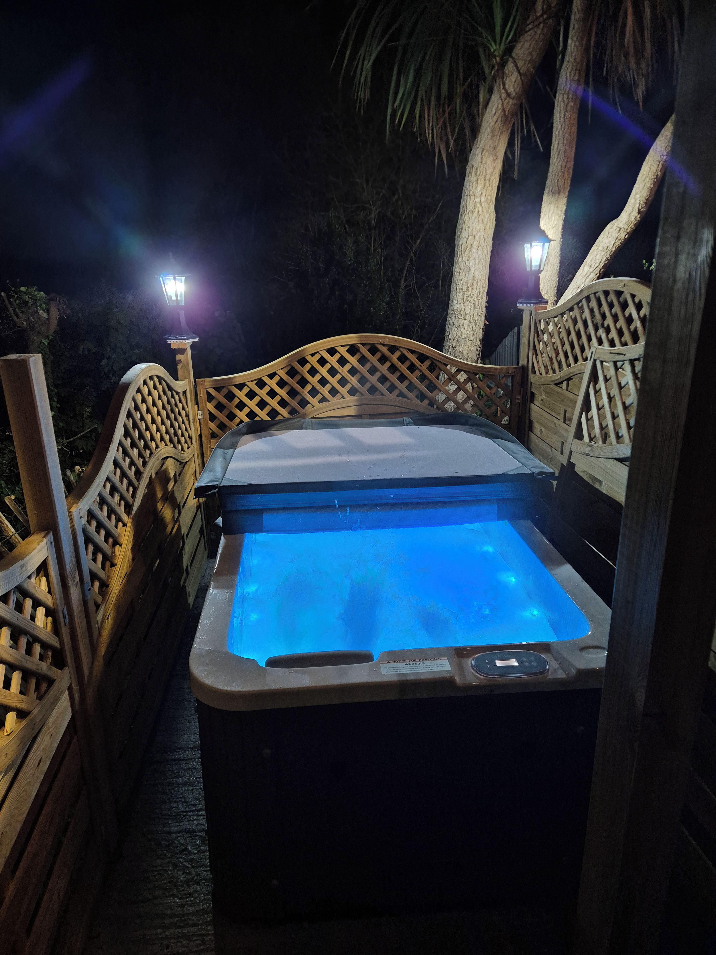 A hot tub with a wooden lattice railing, surrounded by trees and a night sky.