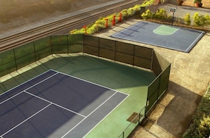 An outdoor tennis court with a green surface, surrounded by a metal fence, and adjacent to a road with a small basketball court. - Carlsbad | Family Beach Adventure | Pool + Beach | 2BR (Carlsbad)