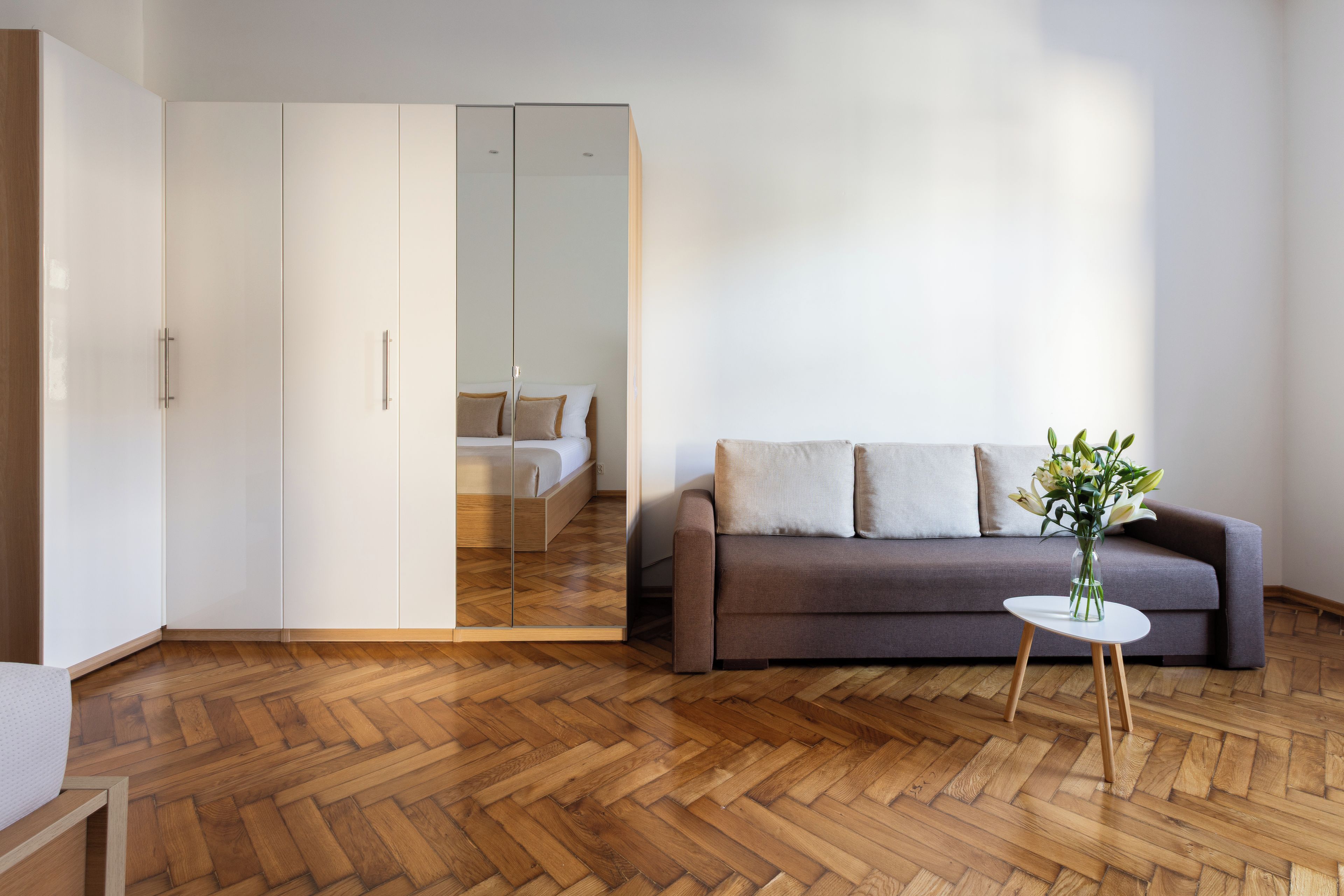 A modern living room with a herringbone wooden floor, a grey sofa with cushions, a small white table with a vase, and a mirrored wardrobe.