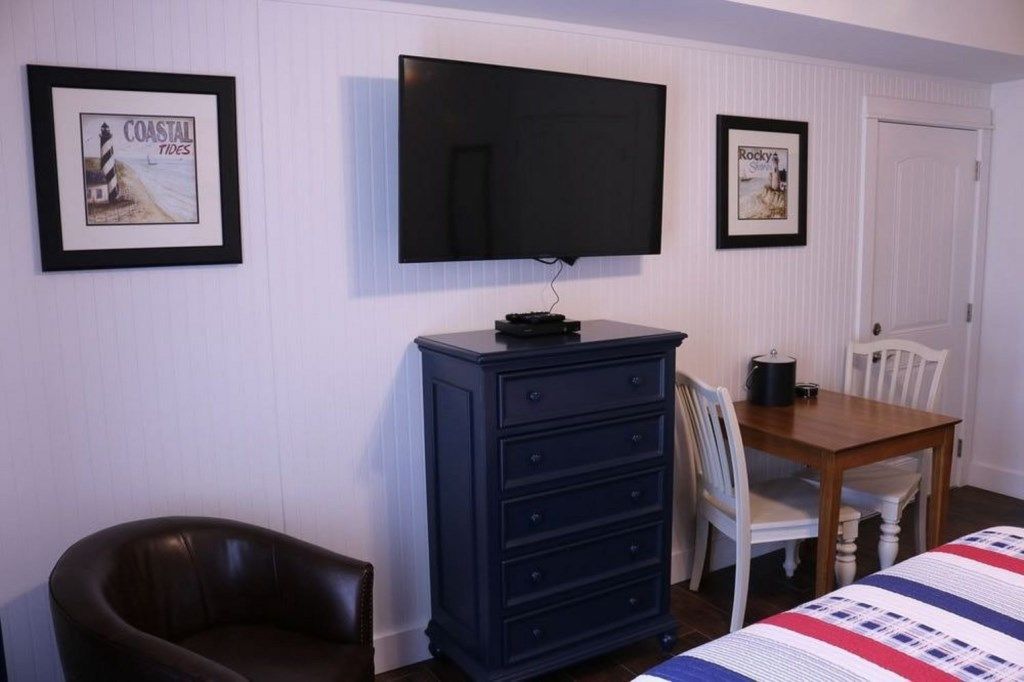 A room with a television mounted on the wall, a dresser, a wooden table with chairs, and framed pictures on the wall.