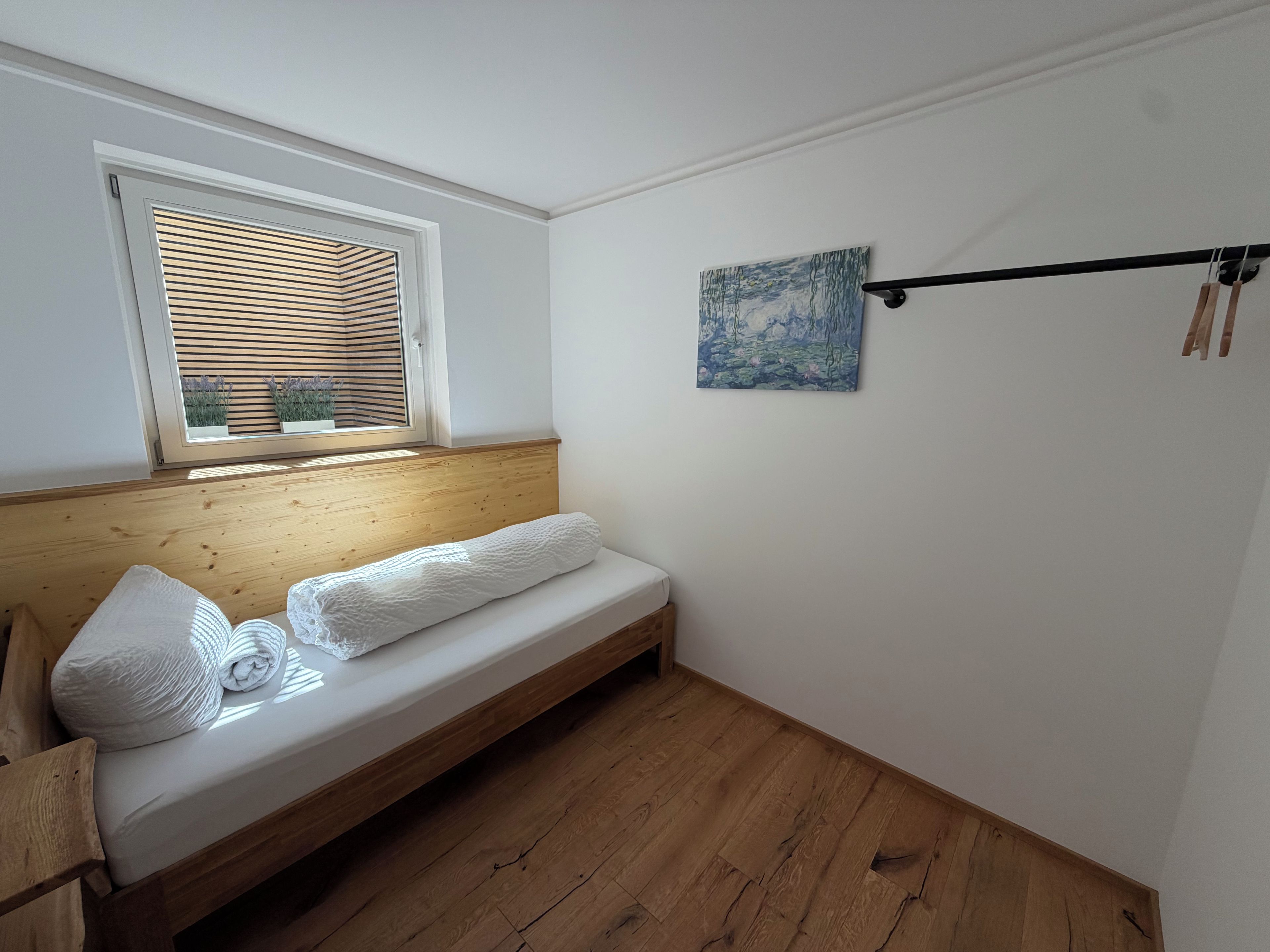 Una habitación pequeña con un banco de madera, una ventana con persianas, un cuadro en la pared y un estante negro fijado al techo.