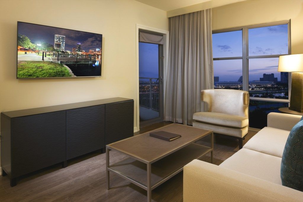A hotel room with a large window offering a city view, a flat-screen TV mounted on the wall, a dark-colored console table, and a beige armchair.