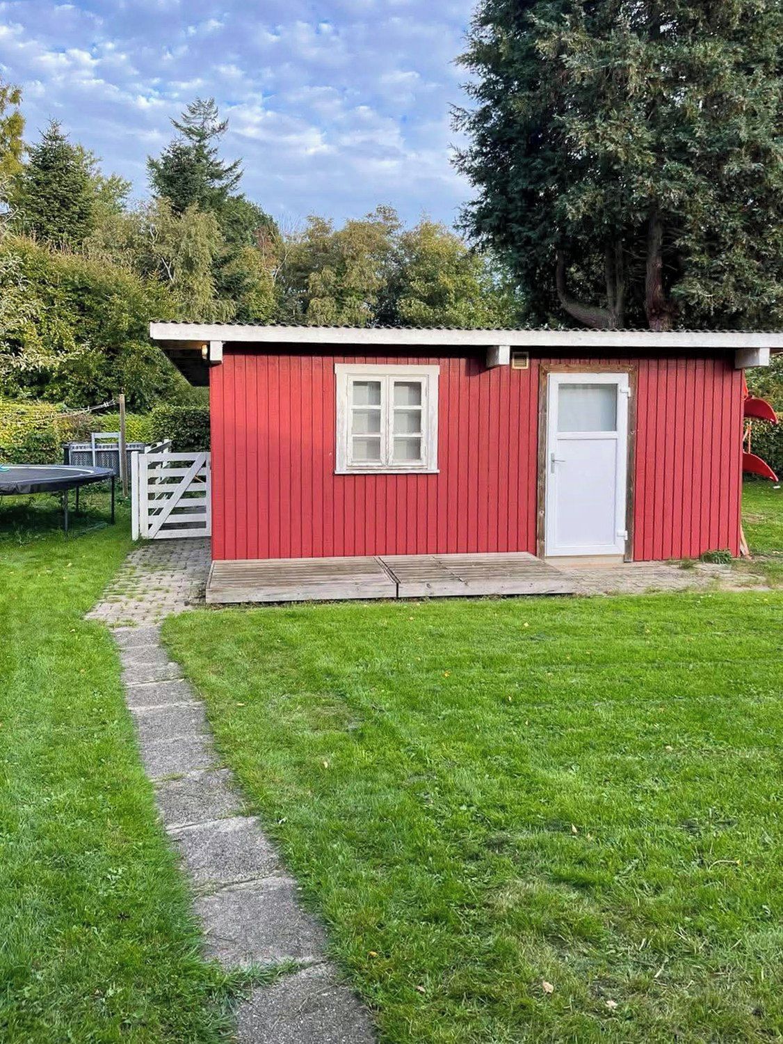 A small red building with a white door and window, a paved path leading to it, and a white gate.