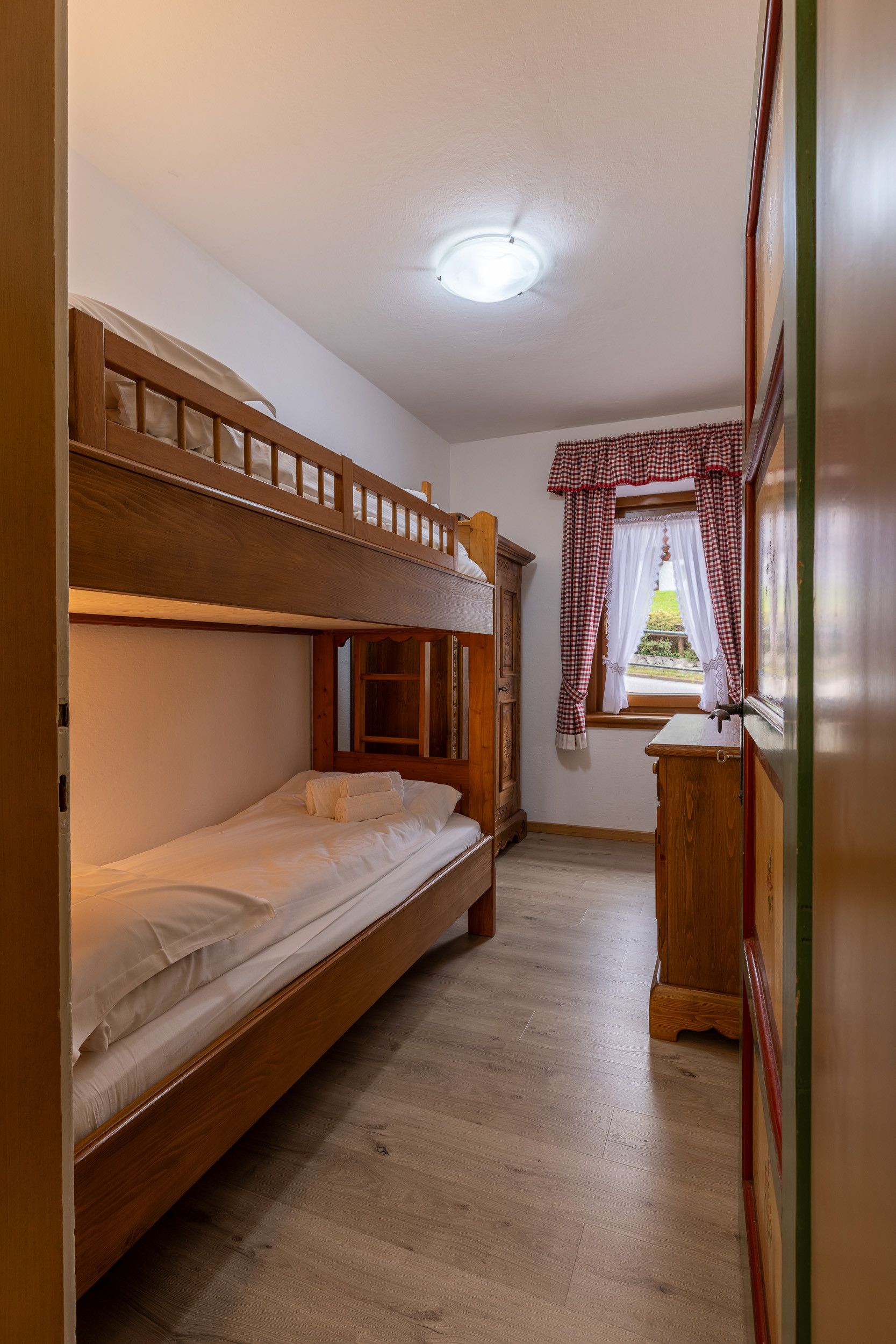 Une chambre avec un lit superposé, une tête de lit en bois, une petite armoire en bois et une fenêtre avec des rideaux à carreaux.