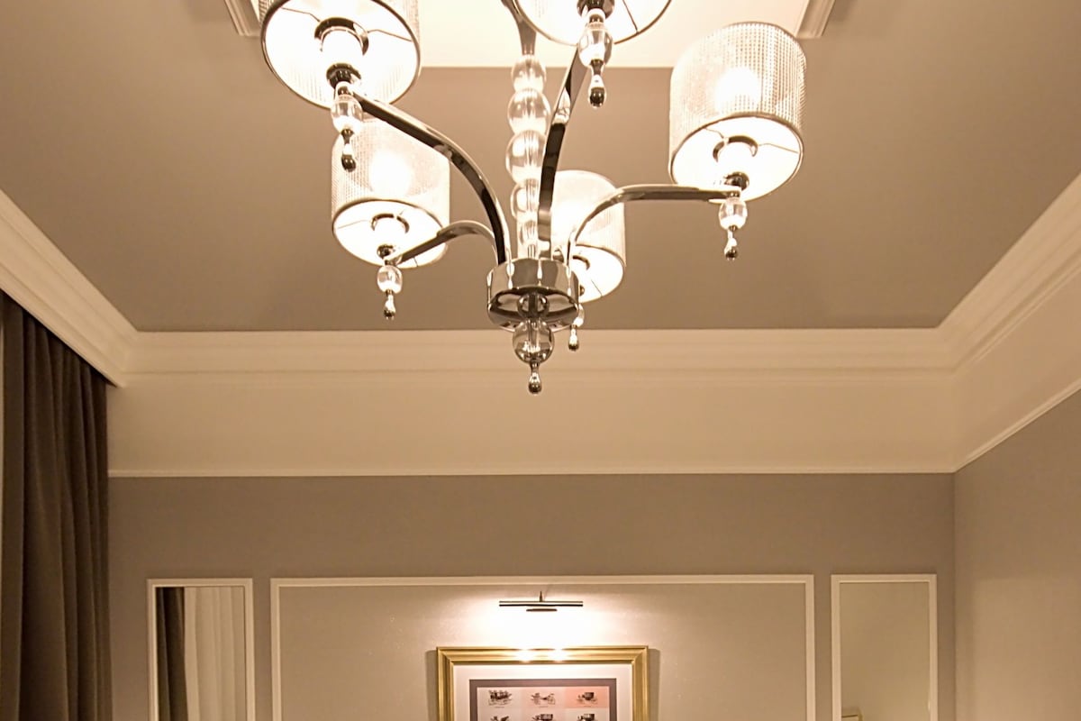 A hotel room with a large bed, a chandelier, two bedside tables with lamps, a framed picture on the wall, and a mirror.