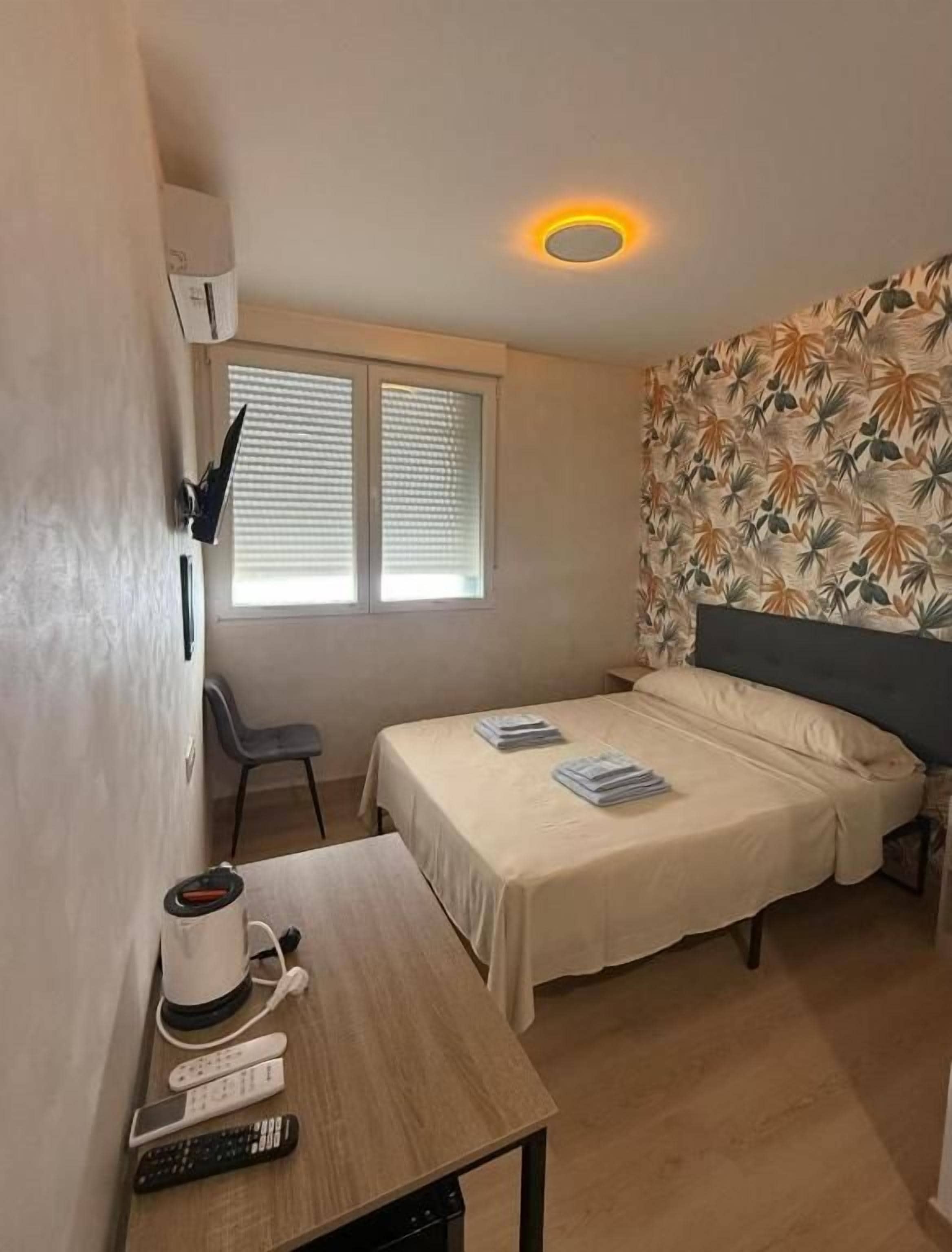 A small room with a bed, a desk with a coffee maker and remote controls, a window with blinds, and a wall with a leaf pattern.