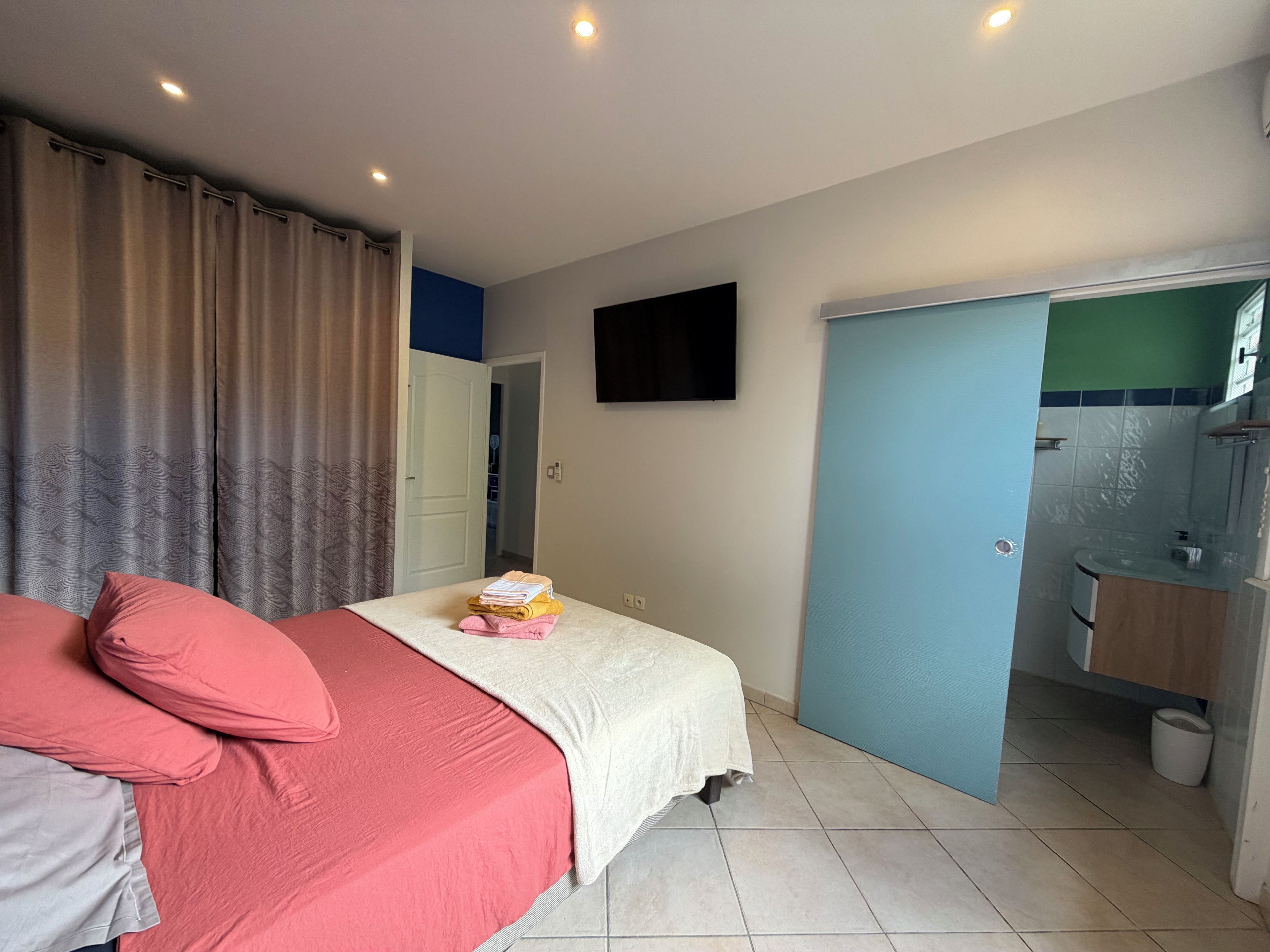 A bedroom with a bed, pillows, a bedside table with books, a TV on the wall, and a bathroom visible through an open door.
