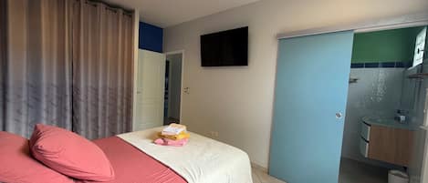 A bedroom with a bed, pillows, a bedside table with books, a TV on the wall, and a bathroom visible through an open door.