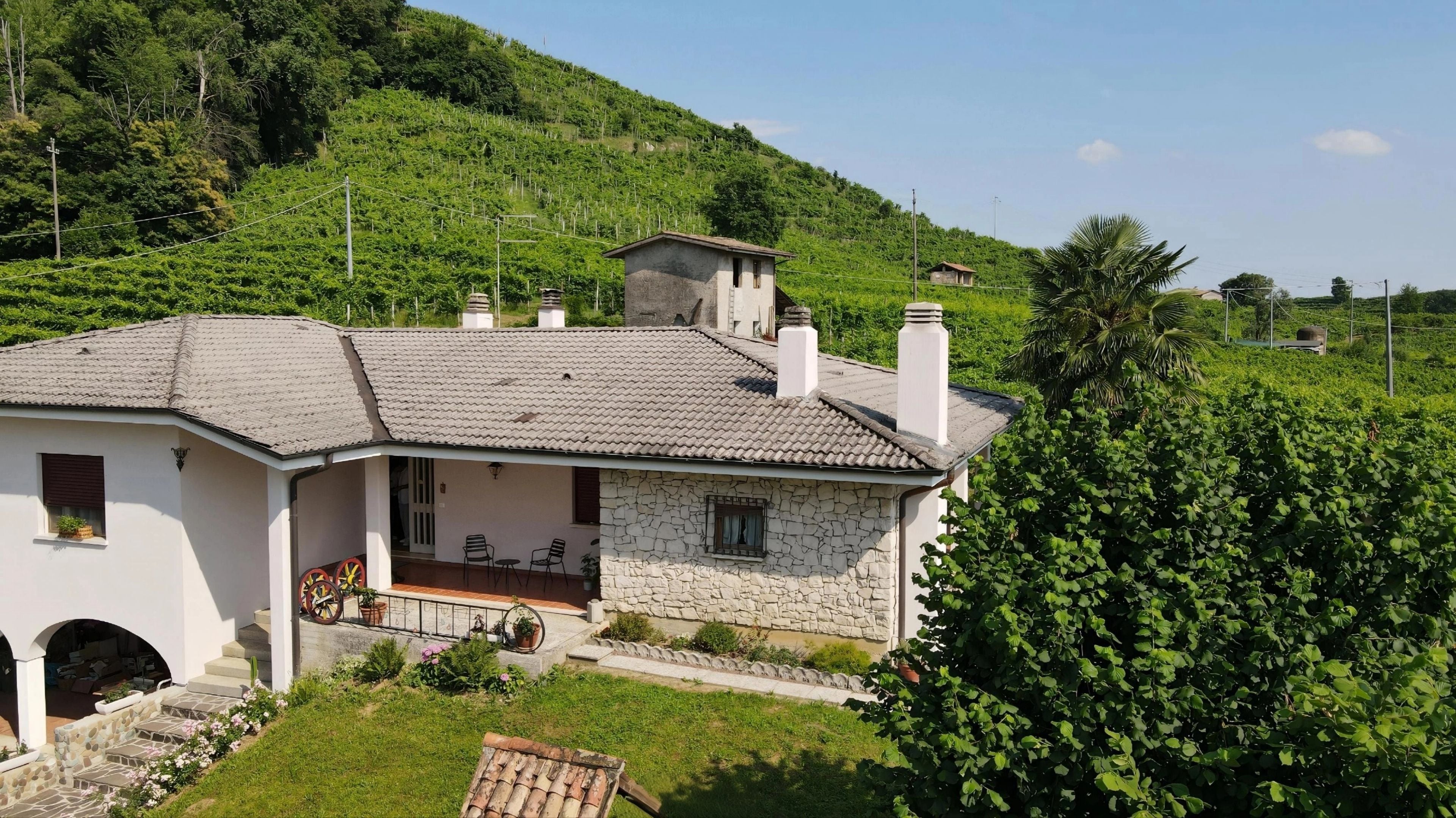 A two-story stone house with a tiled roof, a porch with chairs, and a small garden area.