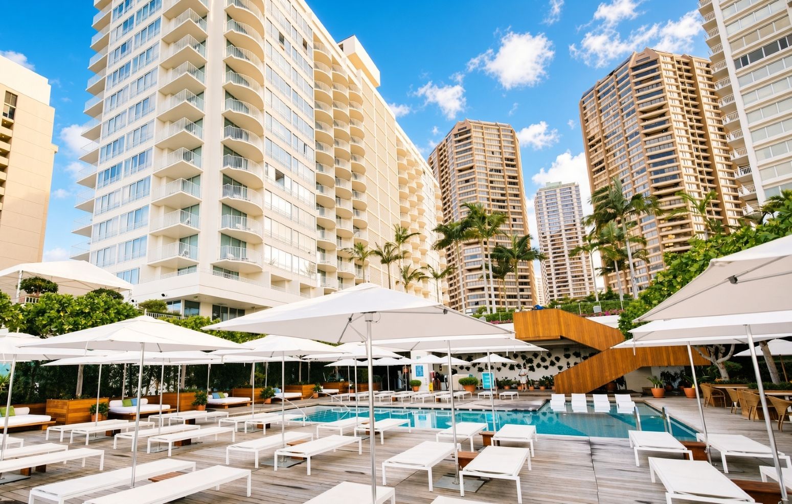 Un espace piscine d’hôtel avec des chaises longues et des parasols, entouré de grands immeubles.