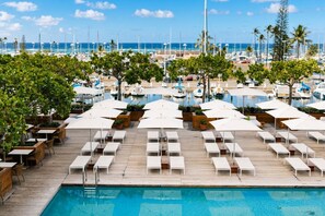 Zona de piscina con sillones de descanso y sombrillas, una terraza de madera y vistas al puerto deportivo con barcos.