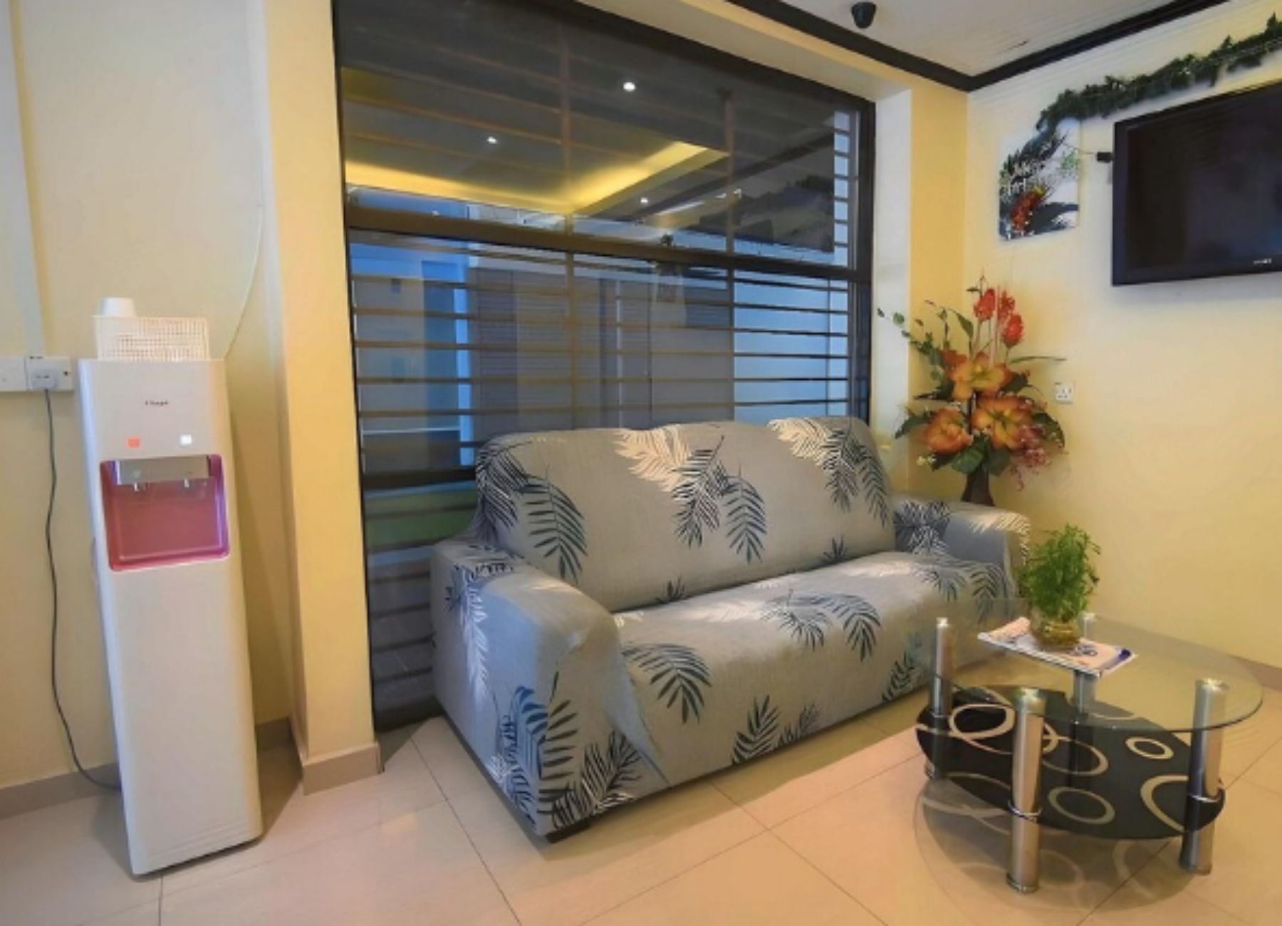 A living room with a sofa, a small table, a water dispenser, and a TV mounted on the wall.