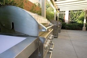 An outdoor grill area with a covered patio, a concrete floor, and surrounding greenery. - Anaheim | Family Theme Park Stay | Pool & Hot Tub | 2BR (Anaheim)