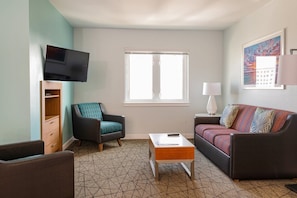 Ein gemütliches Wohnzimmer mit Fernseher, Sofa, Sessel, Couchtisch und Fenster.