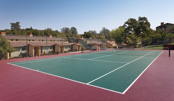 Pista de tennis amb superfície vermella, envoltada per una tanca, i adjacent a una filera d'edificis de fusta.