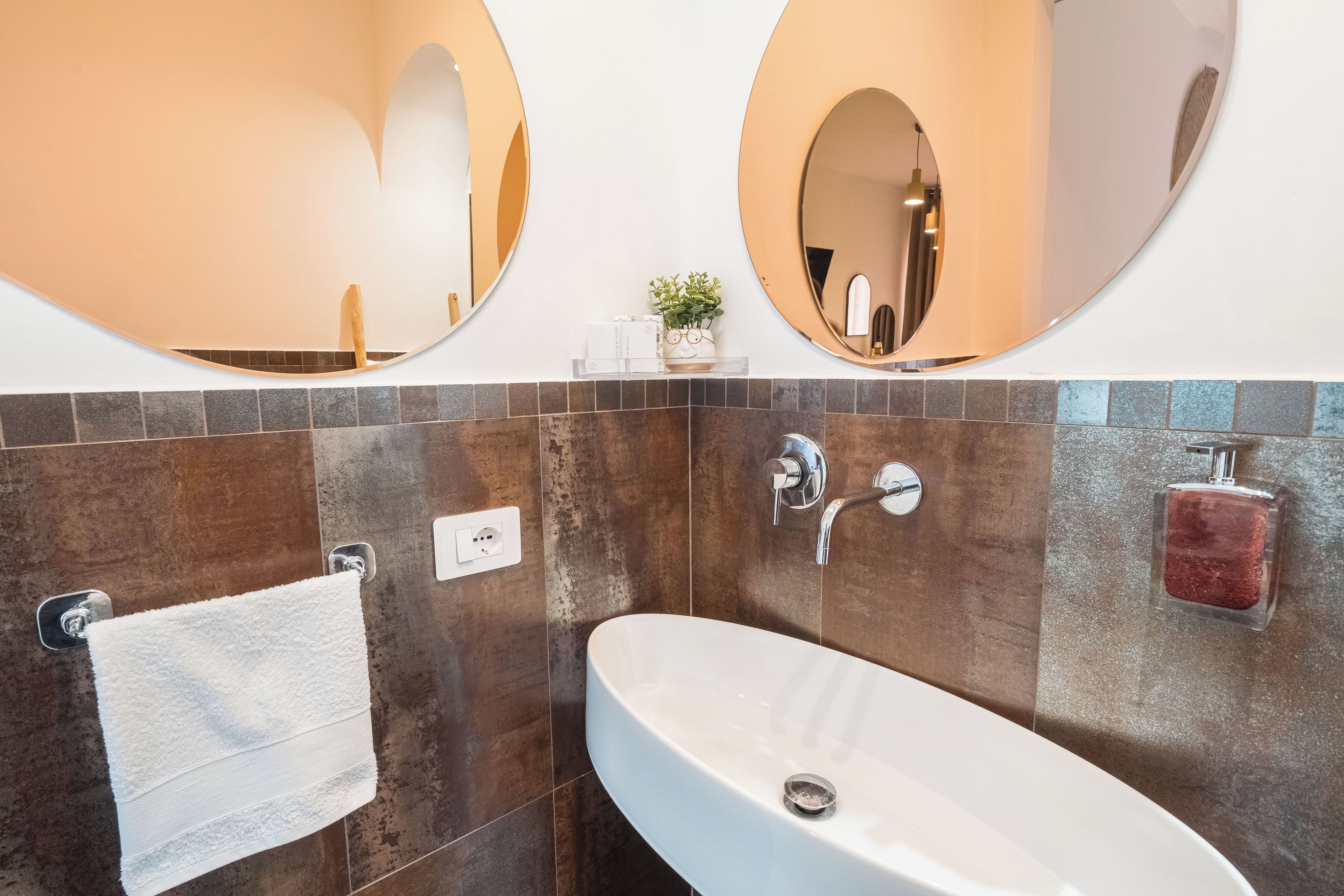 Un baño con un lavamanos blanco, dos espejos, un perchero para toallas y una planta pequeña.