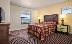 Quarto com uma cama grande, duas mesas de cabeceira com luminárias, uma TV sobre um suporte e uma janela com persianas.