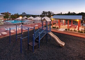 Zona de jocs amb tobogan i elements per escalar, piscina i un pavelló cobert amb bancs.
