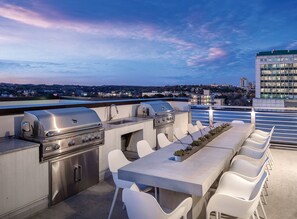 Un patio exterior con una parrilla, asientos y vistas a la ciudad al anochecer.