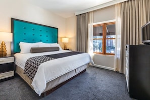 Une chambre d’hôtel avec un grand lit, une tête de lit de couleur turquoise, une fenêtre avec des rideaux, un téléviseur et une table de chevet avec une lampe.