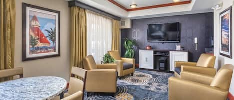A hotel room with a round marble table, beige armchairs, a flat-screen TV, a minibar, and a large window with curtains.