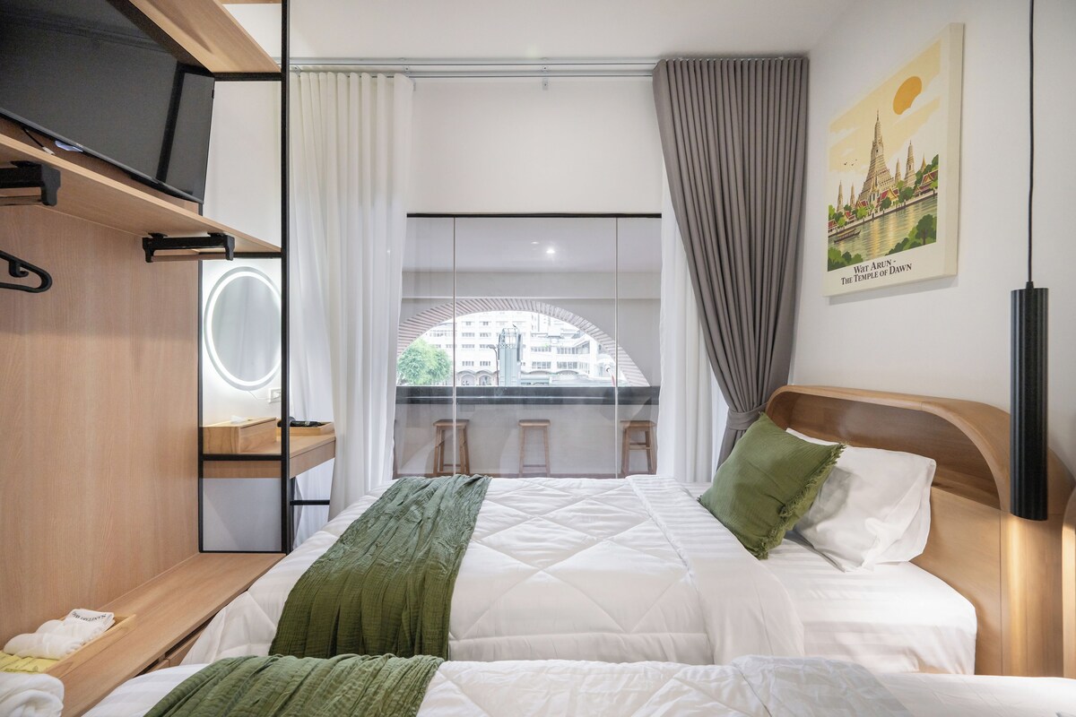 Habitación de hotel con cama, espejo, estante y vista a un edificio desde la ventana.